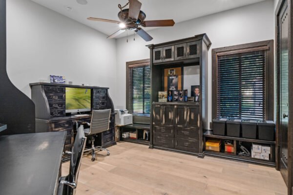 Home office featuring a stylish black desk, modern computer setup, dark wood cabinetry, and large windows with blinds, emphasizing luxury and functionality in the custom estate at 11019 Bridge House Road, Windermere, FL.