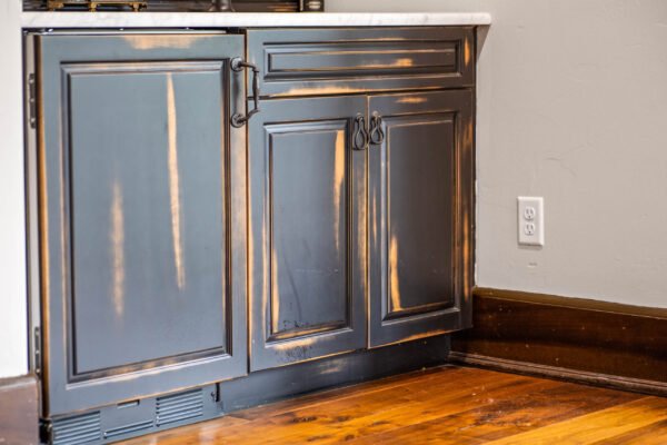 Black kitchen cabinetry with distressed finish, featuring elegant hardware, adjacent to a marble countertop and hardwood flooring, showcasing modern luxury design elements in a high-end home.