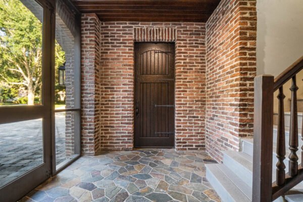 Elegant entryway featuring a rustic wooden door framed by exposed brick walls, complemented by a natural stone floor and wooden staircase, showcasing the luxurious design of the Windermere estate at 11019 Bridge House Road.