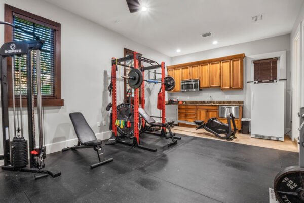 Home gym featuring weightlifting equipment, including a squat rack and bench, alongside a kitchen area with wooden cabinets, emphasizing luxury fitness amenities in the custom estate at 11019 Bridge House Road, Windermere, FL.