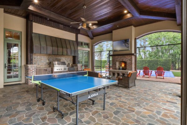 Luxury outdoor entertainment area featuring a blue ping pong table, stone flooring, outdoor kitchen with grill, cozy seating around a fireplace, and a view of the pool in a high-end Windermere estate.