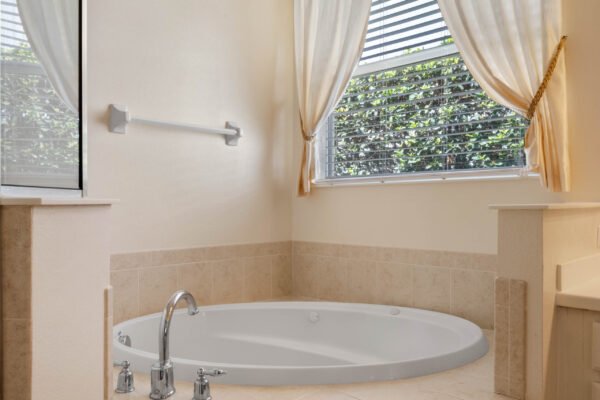 Modern bathroom featuring a spacious round soaking tub, natural light from window with blinds, and elegant drapery, highlighting a relaxing atmosphere in the remodeled home at 15061 Spinnaker Cove Lane, Winter Garden, FL.