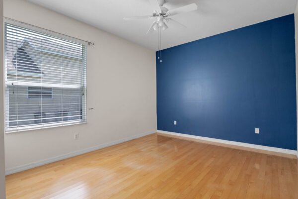 Empty room featuring light hardwood flooring, a blue accent wall, ceiling fan, and window with blinds, suitable for showcasing potential living space in a real estate listing.