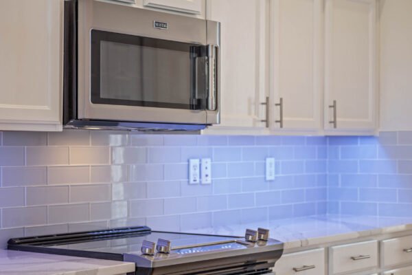 Modern kitchen featuring stainless steel microwave above a sleek black stove, with white cabinetry and gray subway tile backsplash illuminated by under-cabinet lighting.