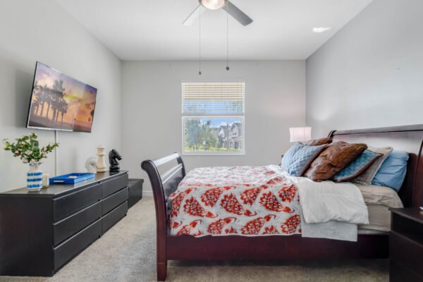 Bedroom with a king-size bed featuring a floral comforter, dresser with a television, and a window showcasing views of the neighborhood, highlighting modern decor and natural light.