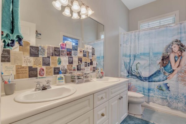 Bathroom featuring dual sinks, a large mirror, and a decorative shower curtain with a mermaid design, surrounded by colorful notes on the wall, emphasizing a personalized and whimsical decor style.