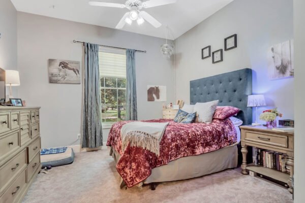 Cozy bedroom featuring a tufted blue headboard, plush bedding in shades of burgundy and gray, decorative pillows, and a nightstand with a lamp, complemented by large windows with blue curtains and framed artwork on the walls.