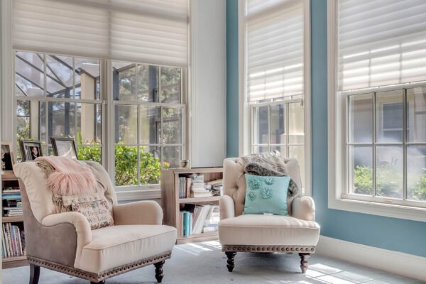 Cozy sunlit sitting area featuring two elegant armchairs, one with a pink throw blanket and decorative pillow, beside a bookshelf, framed by large windows with blinds, showcasing a view of greenery.