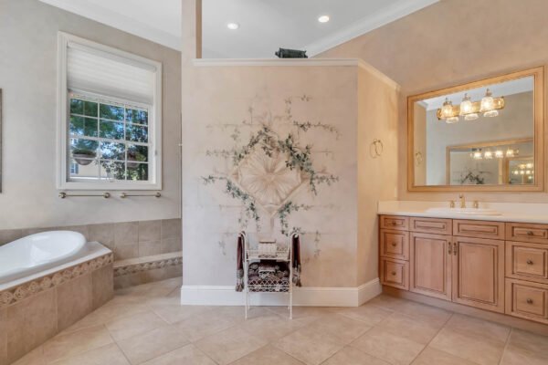 Luxurious bathroom featuring a freestanding soaking tub, elegant cabinetry, decorative floral wall art, and a large mirror with stylish lighting, showcasing the upscale design of the Windermere estate at 6264 Blakeford Drive.