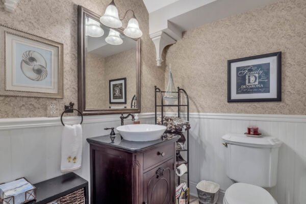 Elegant bathroom featuring a dark wood vanity with a vessel sink, decorative mirror, wall-mounted shelving, and stylish light fixtures, complemented by cream-colored walls and white wainscoting.