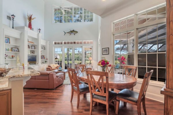 Elegant dining area with wooden table, surrounded by chairs, featuring large windows showcasing views of the manicured landscape, complemented by a cozy living space with a leather couch and decorative elements, in a custom-built estate on Blakeford Drive, Windermere.