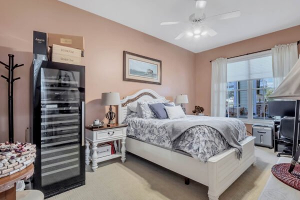 Cozy bedroom featuring a white bed with decorative pillows, a wine cooler beside a nightstand, and a window with natural light illuminating the space, reflecting a comfortable living environment in Windermere, FL.