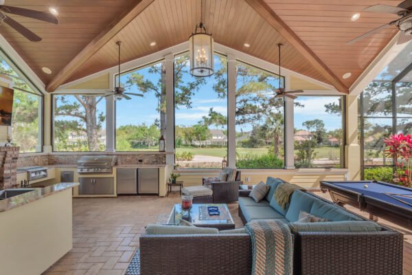 Luxurious indoor-outdoor living space featuring a spacious lounge area with modern furniture, large windows overlooking the golf course, and a well-equipped outdoor kitchen, ideal for entertaining in the custom-built estate at 6264 Blakeford Drive, Windermere, FL.