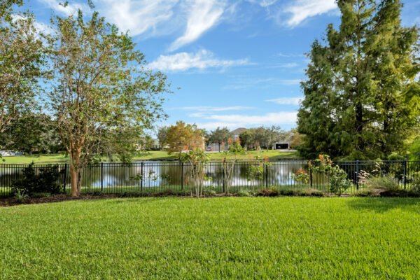 Backyard view of 13230 Fossick Road in Windermere, showcasing lush green lawn, ornamental trees, and a serene pond with a black fence, emphasizing the home's premium pond lot location.