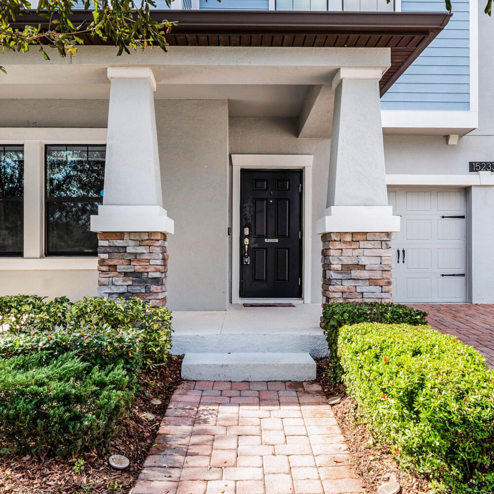 Front entrance of 15233 Night Heron Drive, Winter Garden, FL, featuring a black door, stone-accented columns, landscaped shrubs, and a brick paver walkway.