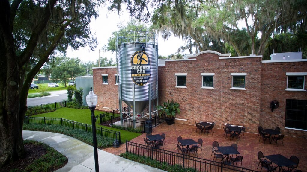 Crooked Can Brewing Company exterior with large silver fermentation tank, brick building, outdoor seating area, and lush greenery in Winter Garden, Florida.