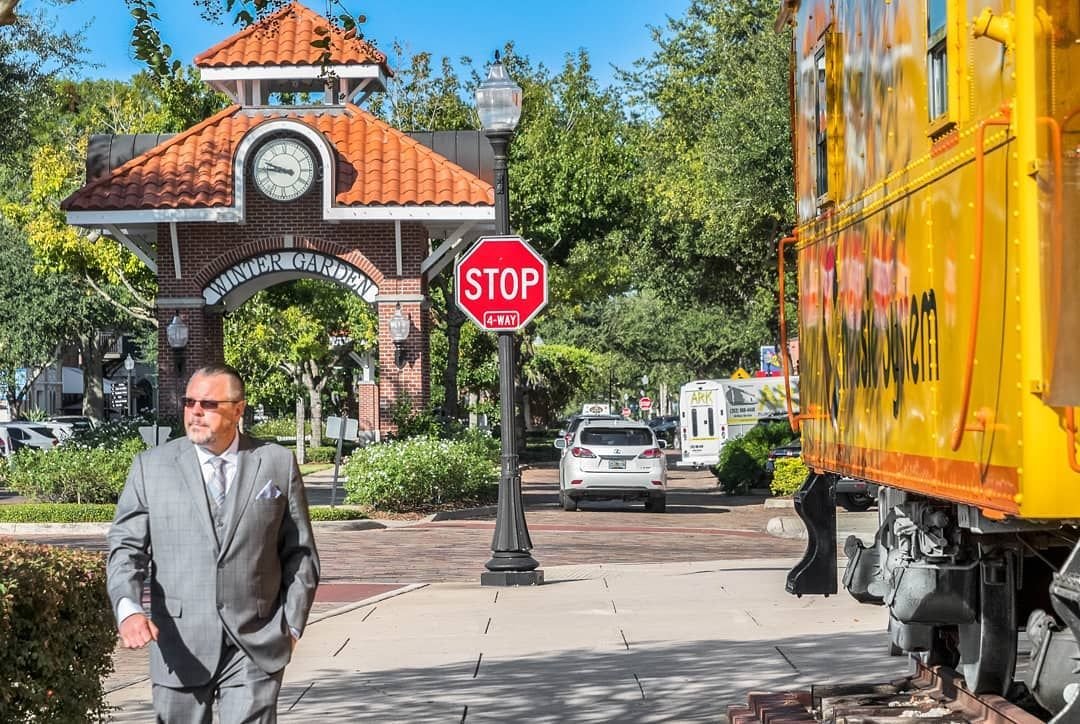 Man in a suit walking past the Winter Garden entrance, with a stop sign and a vintage train car in view, representing real estate activity in Orlando.
